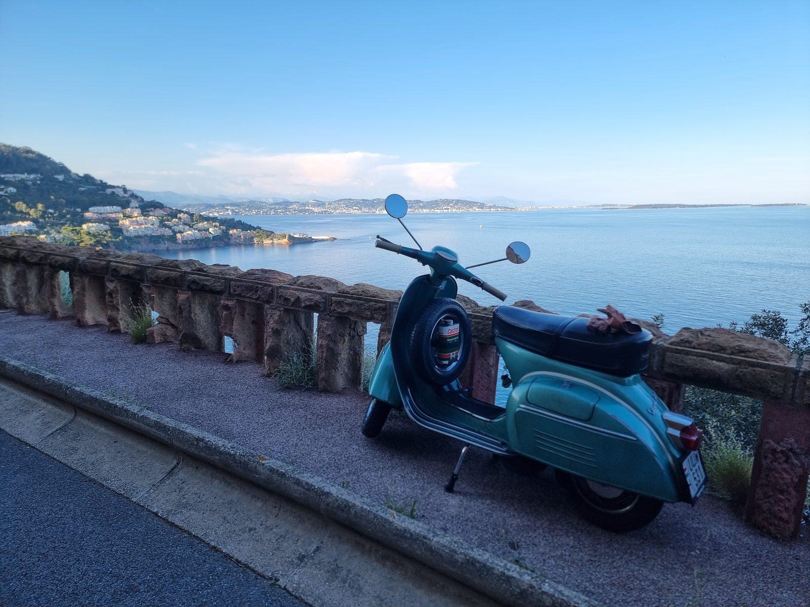 Türkizzöld klasszikus Vespa egy tengerparti kilátó kőkorlátjánál, háttérben a mediterrán öböl és a dombok